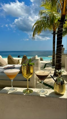 three cocktails sitting on top of a table next to the ocean and palm trees