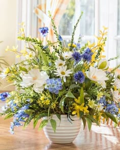 a white vase filled with blue and yellow flowers on top of a wooden table next to a window