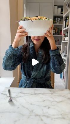 a woman sitting at a kitchen table holding up a bowl with food in front of her face