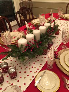 the table is set for christmas dinner with red and gold place settings, silverware, candlesticks and greenery