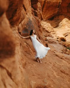 a woman in a white dress is standing on the side of a rock formation with her arms outstretched