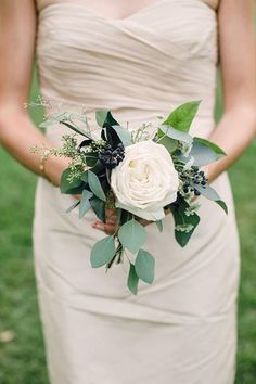 a woman in a dress holding a white rose and greenery bridally bouquet