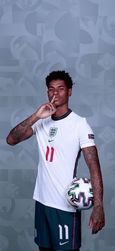 a man with tattoos holding a soccer ball and finger to his mouth while standing in front of a wall