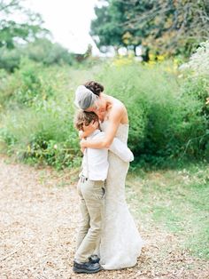 a woman in a wedding dress hugging a young boy on the side of a dirt road