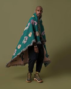 a man wearing a green shawl with flowers on it, standing in front of a brown background