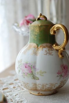 a white and green vase sitting on top of a lace covered tablecloth next to pink flowers