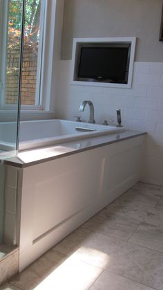 a bath tub sitting under a window next to a tv mounted on the side of a wall