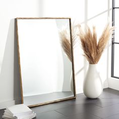 a white vase with some dry grass in it next to a mirror on the floor