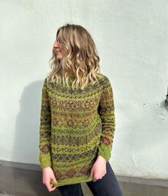 a woman standing in front of a white wall wearing a green sweater and black jeans