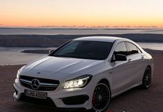 a white mercedes cla parked in front of the ocean at sunset with mountains in the background