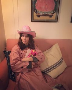 a woman wearing a pink hat and trench coat sitting on a couch with her arms crossed