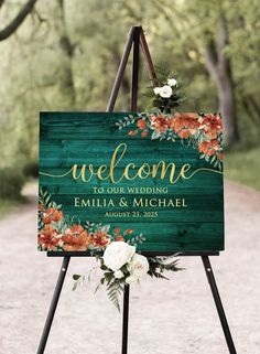 a welcome sign with flowers and greenery sits on an easel in the middle of a dirt road