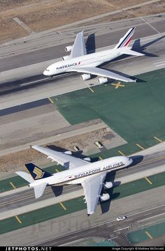 two airplanes are flying side by side in the air over an airport runway and parking lot