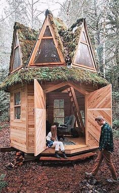 two people are sitting on the porch of a tiny cabin in the woods with moss growing on top