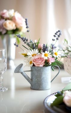 A small metal watering can, perfect for baby shower centerpieces, holds a bouquet of pink roses, white daisies, and lavender sprigs. It's placed on a glossy surface next to a wine glass, with blurred floral arrangements in the background. Watering Can Centerpiece Wedding, Baby In Bloom Table Centerpieces, Baby In Bloom Baby Shower Centerpieces, A Baby In Bloom Baby Shower Ideas, Spring Floral Baby Shower Ideas, Flower Garden Baby Shower Theme, April Showers Bring May Flowers Baby, Spring Girl Baby Shower Ideas, Baby Shower Bloom Theme