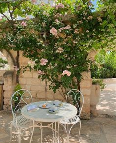 an outdoor table and chairs under a tree with pink flowers on the top, in front of a stone wall