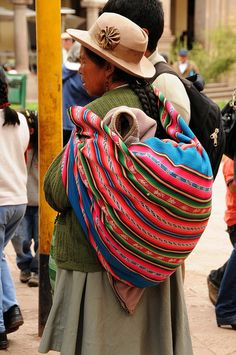 a woman carrying a baby in a colorful bag on her back while walking down the street