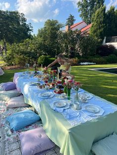 a woman setting up a long table with plates and glasses on it in the grass