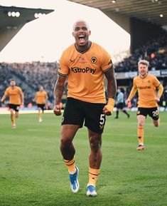 a man in an orange shirt and black shorts is running on the soccer field with his mouth open