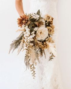 a bride holding a bouquet of flowers and greenery on her wedding day in front of a white wall