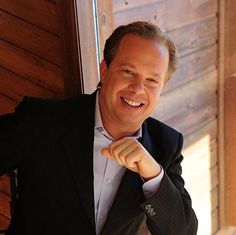 a man in a suit is smiling and pointing his finger at the camera while leaning against a wooden wall