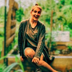 a woman sitting on top of a wooden bench next to a green plant filled garden