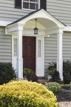 a house with white trim and brown front door on the side of it, surrounded by shrubbery