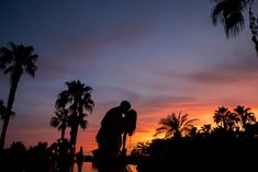 two people standing next to each other in front of palm trees and water at sunset