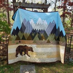 a bear quilt hanging on a fence in the woods with mountains and trees behind it