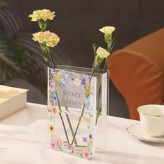 three yellow flowers in a clear vase sitting on a table next to a cup and saucer