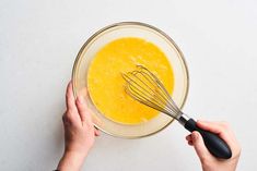 two hands holding a whisk in a glass bowl with yellow liquid on the side