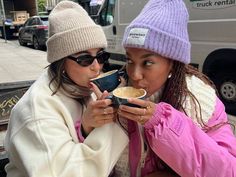 two women sitting on a bench drinking from cups in front of a van and truck