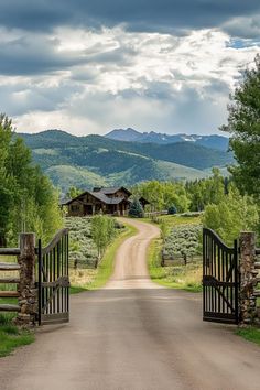 an open gate leading to a dirt road with mountains in the background and trees on either side