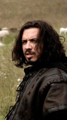 a man with long hair standing in a field next to some sheep and looking at the camera