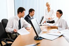 three business people sitting at a table with papers in front of them and one woman standing behind the desk