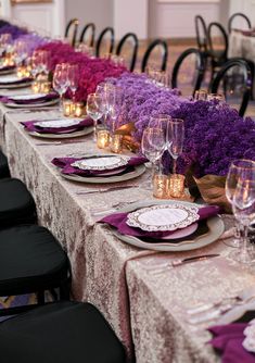 a long table is set with purple flowers and white plates, silverware, and wine glasses