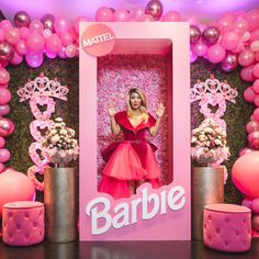 a woman in a pink dress standing behind a barbie photo booth with balloons and decorations