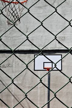 a basketball hoop is seen through a chain link fence on a concrete wall, with a basketball in the foreground