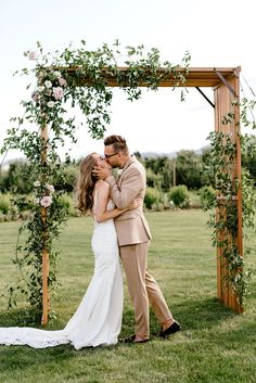 a bride and groom kissing in front of an outdoor wedding ceremony arch with greenery