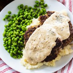 a white plate topped with meat and mashed potatoes covered in gravy next to peas