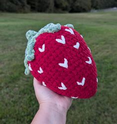 a hand holding up a crocheted strawberry hat in the grass with trees in the background