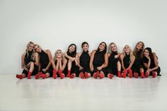 a group of women sitting next to each other in front of a white wall with red shoes