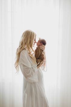 a woman holding a baby in her arms and kissing it's face with the window behind her