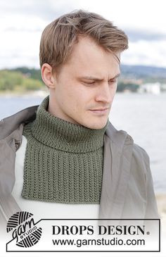 a man wearing a green knitted scarf and jacket looking at his cell phone while standing next to the water