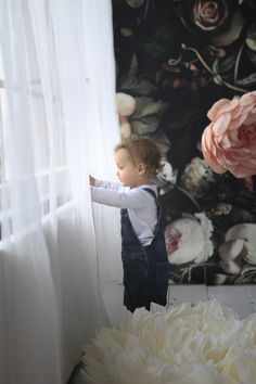 a little boy standing in front of a window with flowers on the wall behind him