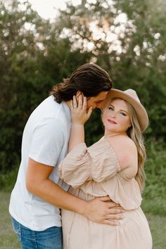 a pregnant woman hugging her husband's chest in an open field with trees in the background