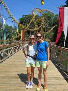 two women standing on a wooden bridge in front of an amusement park roller coaster ride