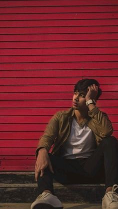 a man sitting against a red wall talking on a cell phone while holding his hand up to his ear