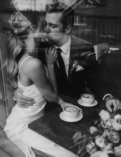 a man and woman kissing while sitting at a table with cups of coffee in front of them