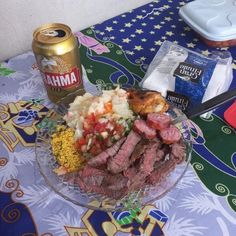 a table topped with a glass bowl filled with meat and veggies next to a can of beer